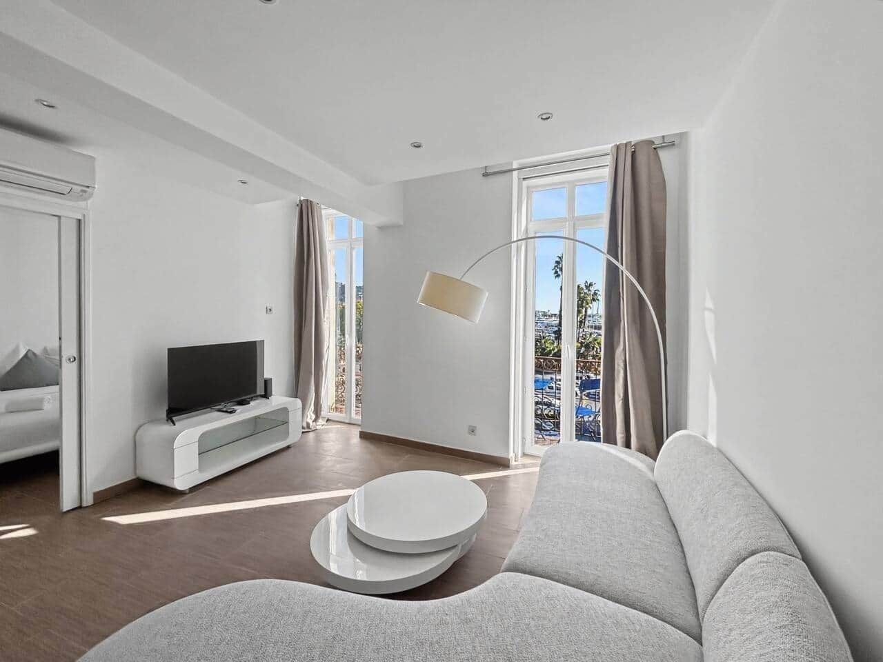 A modern living room with a sofa, a TV on a white console, a floor lamp, and a balcony with a view.