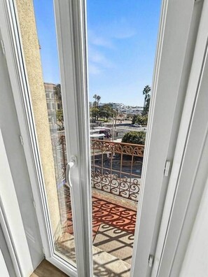 A view through a glass door to a balcony with patterned flooring and a metal railing. - Cannes Port | Suite 1 | Balcon vue Palais (Cannes)