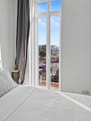 A bedroom with a large window, a bed, a lamp, and a view of the city. - Cannes Port | Suite 1 | Balcon vue Palais (Cannes)