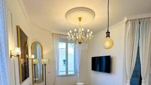 A modern living room with a sofa, a coffee table, a dining table, a chandelier, and a flat-screen TV mounted on the wall. - Cannes Port | Suite 4 | 2 min Palais (Cannes)
