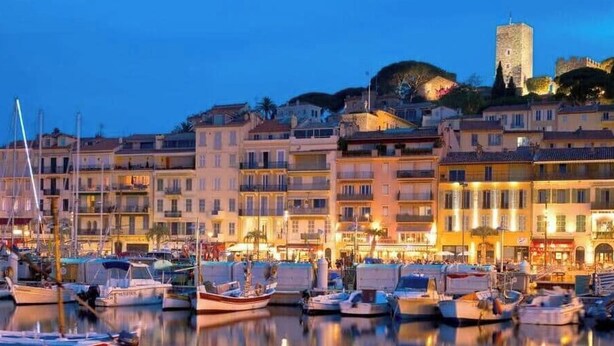 A marina with boats docked, waterfront buildings, and a castle-like structure in the background. - Cannes Port | Suite 4 | 2 min Palais (Cannes)