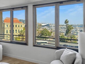 Ein Zimmer mit großen Fenstern, einem weißen Stuhl und Blick auf eine Stadtlandschaft mit Gebäuden und einem Yachthafen.