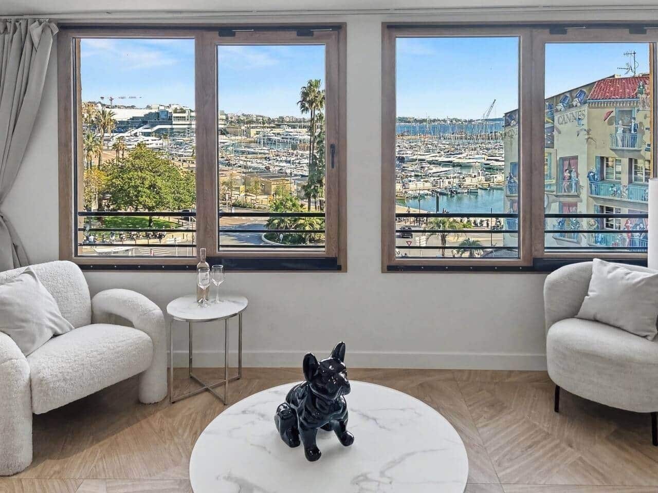 A room with two white armchairs, a small round table with a glass of water, and a black dog statue. Large windows offer a view of a marina and buildings.