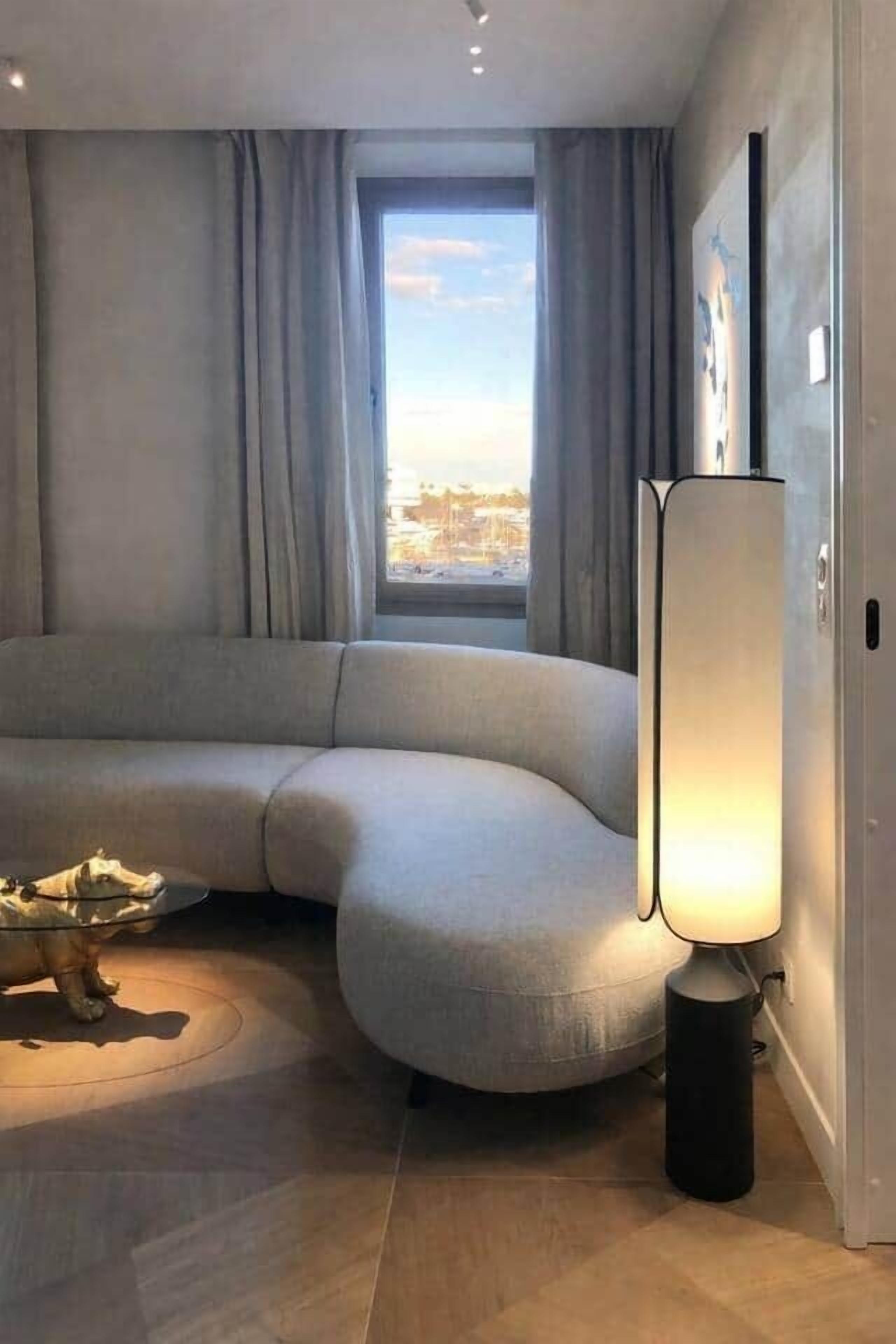 Ein modernes Wohnzimmer mit einer gebogenen Eckcouch, einer Stehlampe und Blick durch ein Fenster.