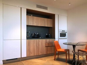 A modern kitchen with wooden cabinets, a built-in oven, and a dining area with two chairs and a glass table. - Cannes Port | Suite 2 | Vue imprenable Palais (Cannes)
