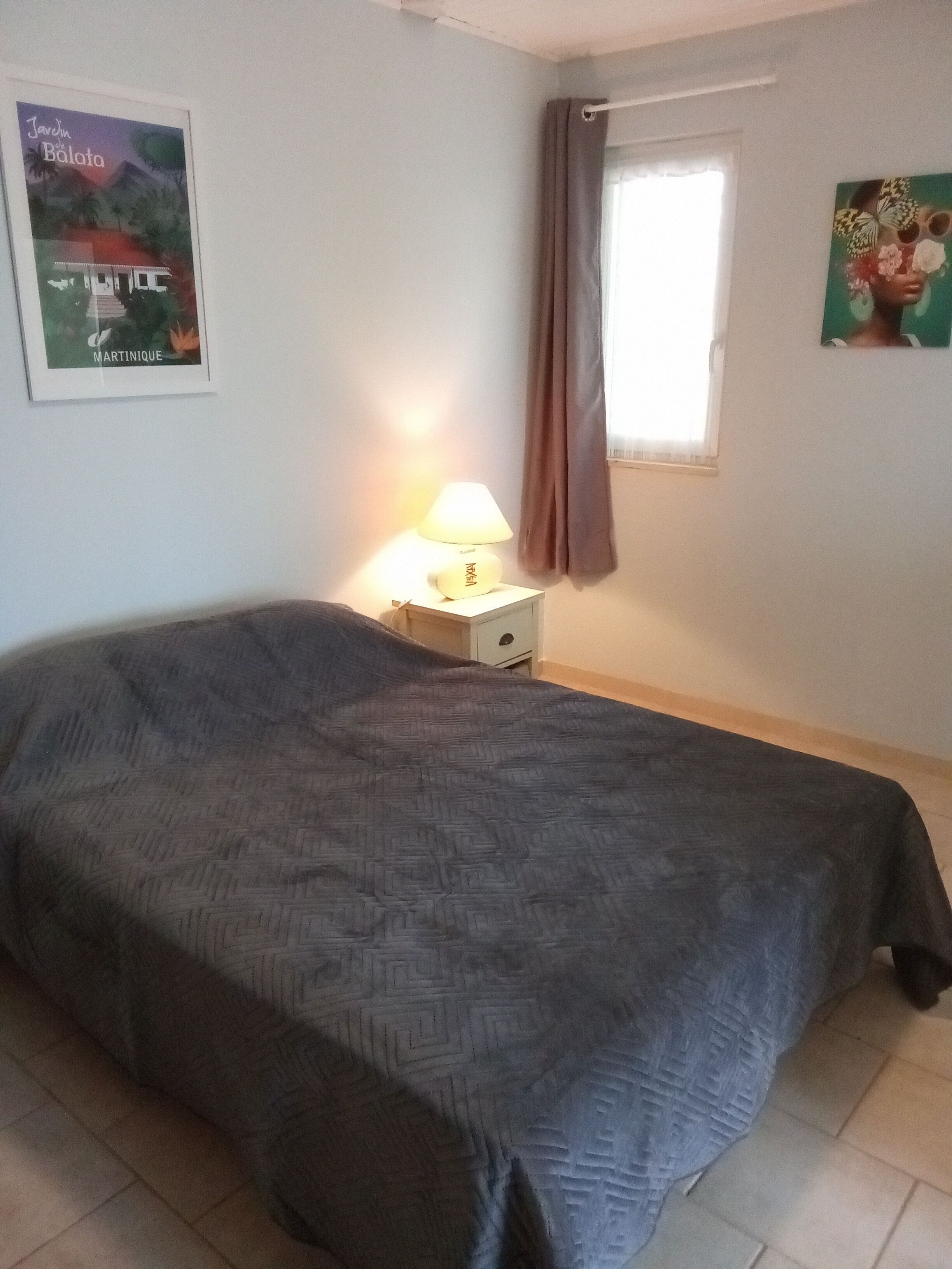 A single bed with a patterned bedspread, a bedside table with a lamp, a poster on the wall, and a window with curtains.