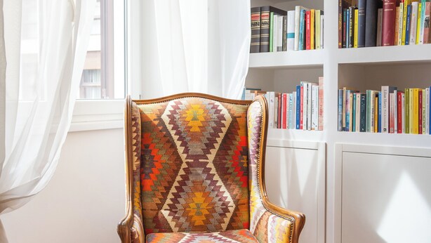 Ein Zimmer mit einem gemusterten Sessel, einem Bücherregal und einem Fenster mit durchsichtigen Vorhängen.