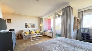 Ett hotellrum med en säng, ett skrivbord med en stol, en soffa med kuddar, ett element och ett fönster med gardiner.