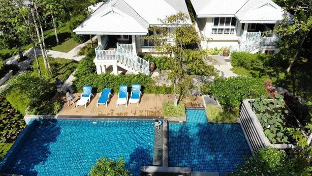 Ein großer Swimmingpool mit Holzterrasse und Liegestühlen, umgeben von üppiger Bepflanzung und zwei weißen Gebäuden mit Balkonen.