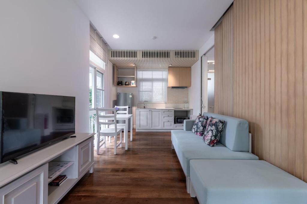 A modern living room with a TV, white furniture, a sofa, and a kitchen in the background.