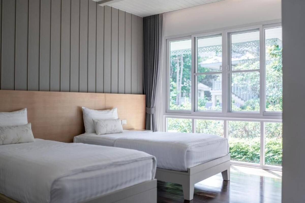 A bedroom with two beds, a large window with a view of greenery, and a wooden headboard.