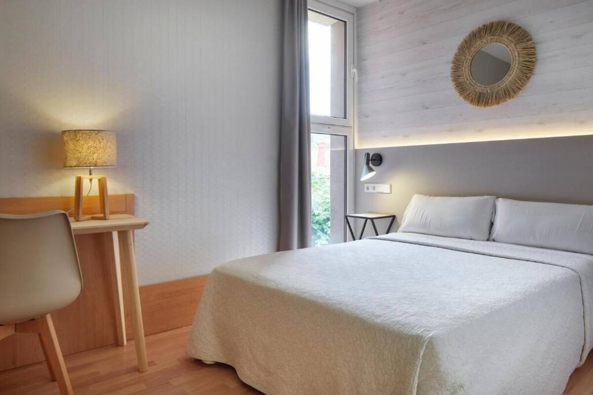 Habitación de hotel con cama, escritorio con lámpara, una mesita y espejo, con vista al exterior.