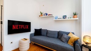 Ett vardagsrum med en grå soffa, en väggmonterad tv som visar ”NETFLIX” och ett litet sidobord med en lampa.