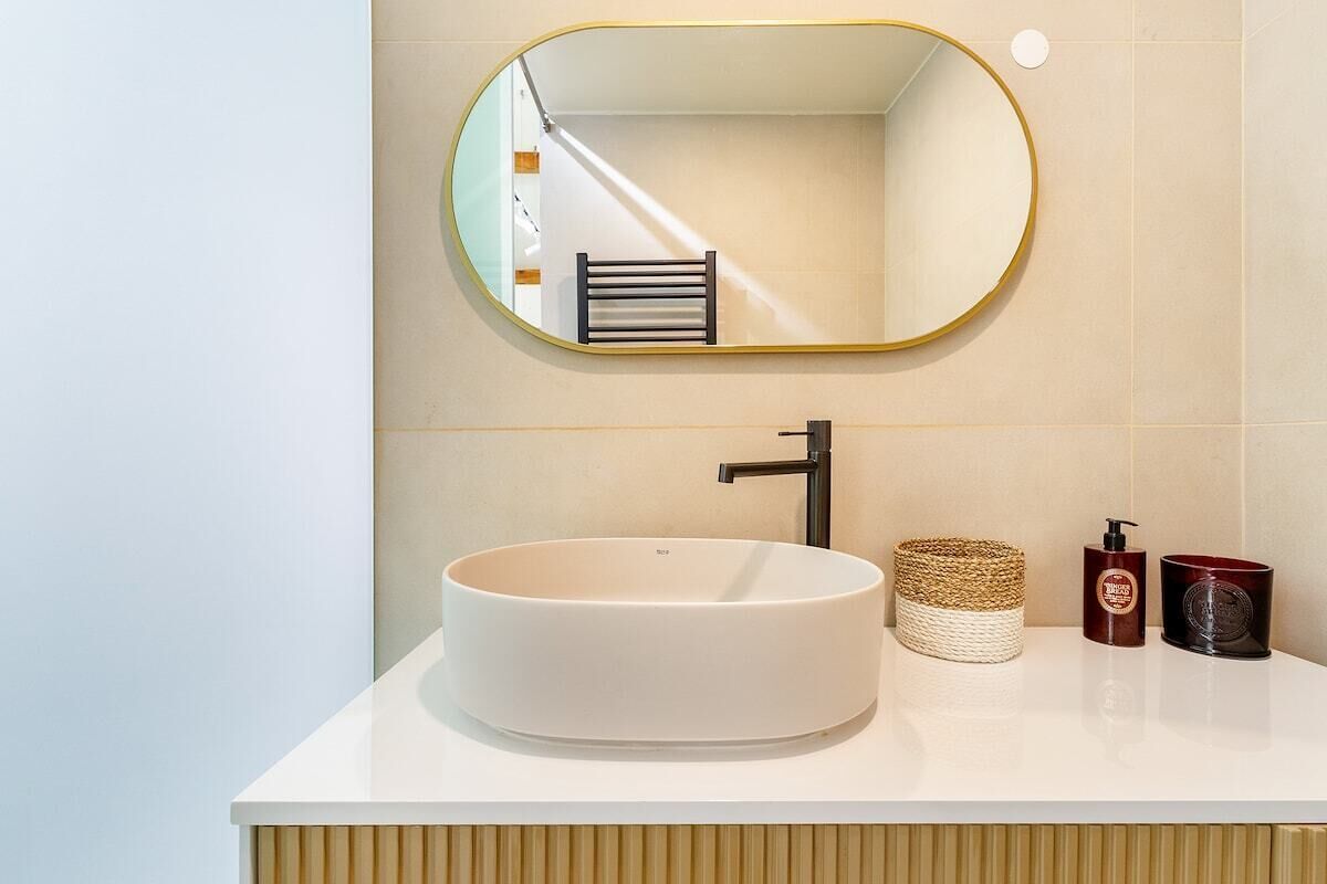 Ein Badezimmer mit einem runden, weißen Waschbecken, einem modernen Wasserhahn, einem Spiegel mit goldener Rahmung und einem Handtuchhalter.