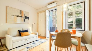 Uma sala de estar moderna com um sofá branco, uma mesa de jantar com cadeiras, uma janela grande com vista para o verde, e um aparelho de ar condicionado na parede.