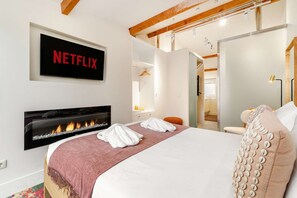 Ein modernes Hotelzimmer mit Kamin, einem Bett mit weißen Leinentextilien, einer rosa Tagesdecke und einem wandmontierten Fernseher, der 'NETFLIX' anzeigt.