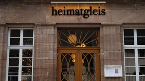 A building with a sign reading 'heimatgleis' above a wooden door with glass panels. There are two windows on either side of the door, and a bench is visible to the right. - heimatgleis (Brilon)