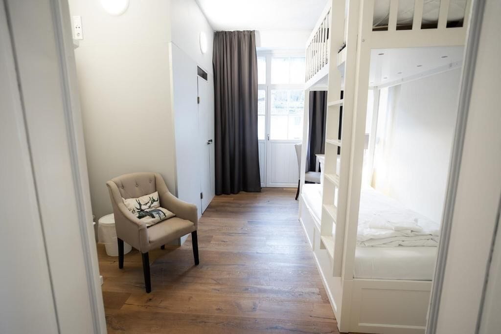A room with a bunk bed, a chair with a cushion, a window with curtains, and a wooden floor.