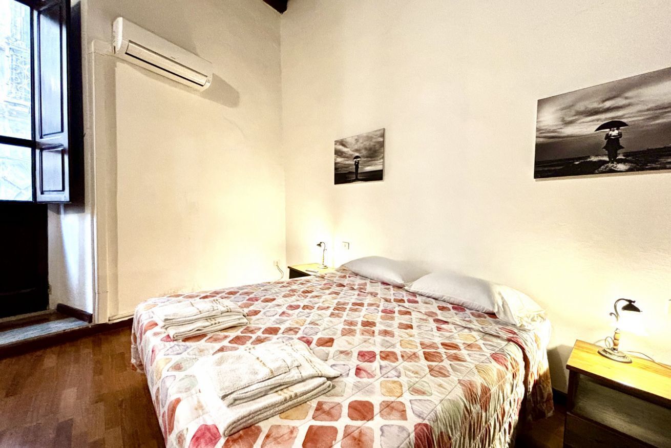 A bedroom with a bed, a nightstand, a lamp, and a wall-mounted air conditioner.