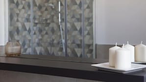 Ein Badezimmer mit einer Dusche, deren Wand ein geometrisches Muster aufweist, einer weißen Kerze auf einer schwarzen Arbeitsplatte und einer Glasdusche.