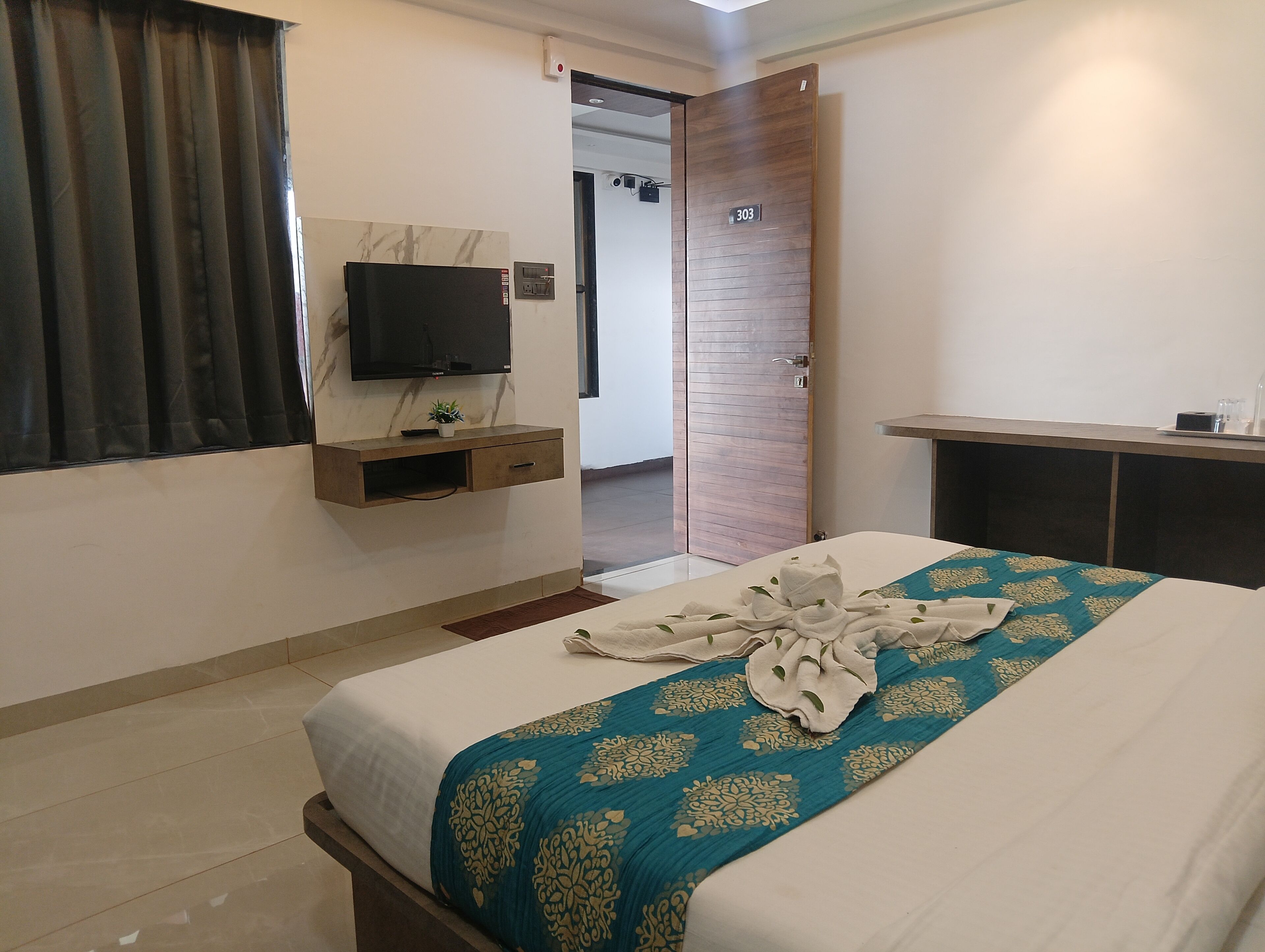A hotel room with a bed, a TV mounted on the wall, a wooden door, and a small desk with a plant.