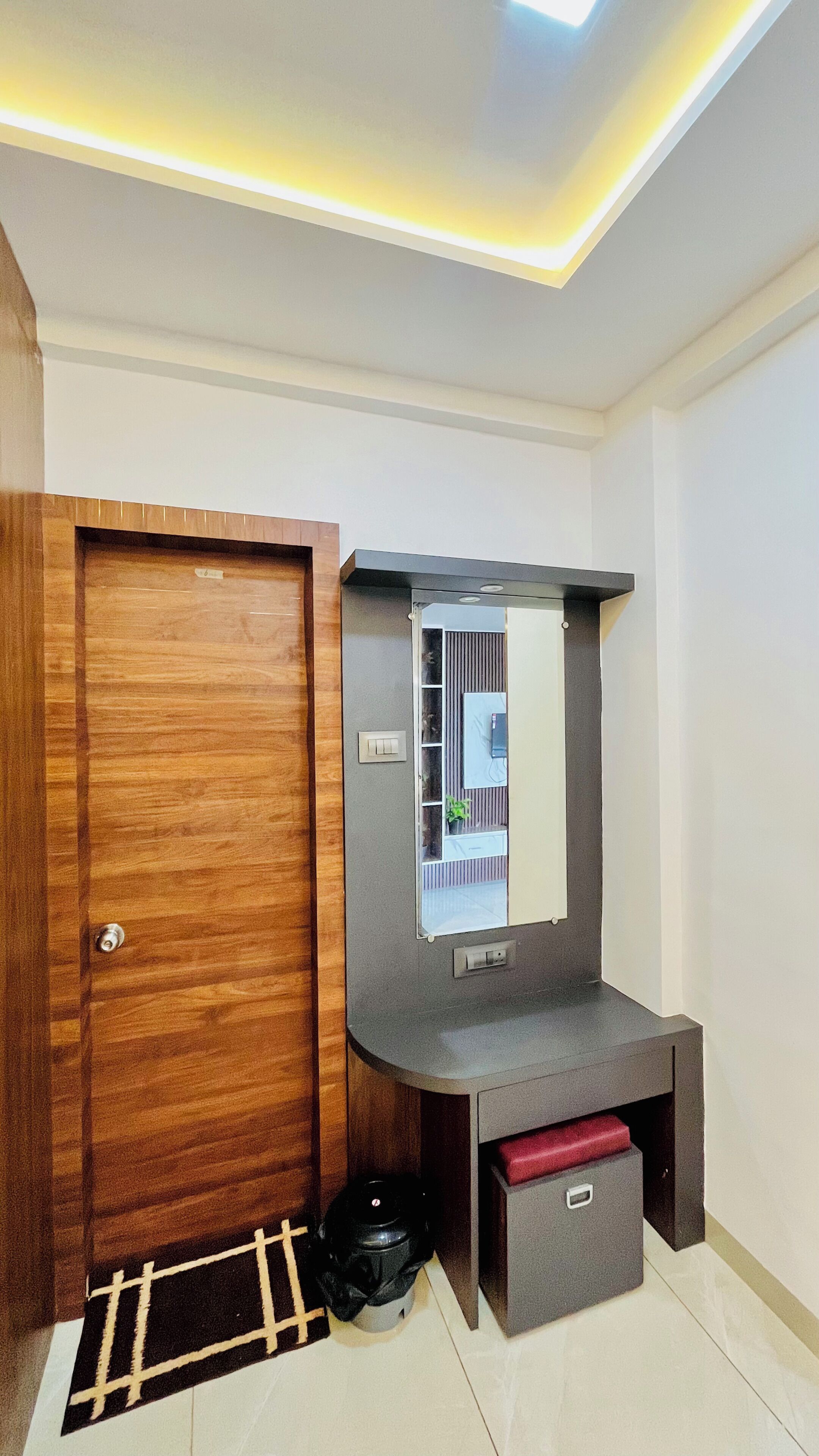 Una habitación compacta con una puerta de madera, un tocador gris con espejo, un pequeño armario rojo y un cubo de basura negro.