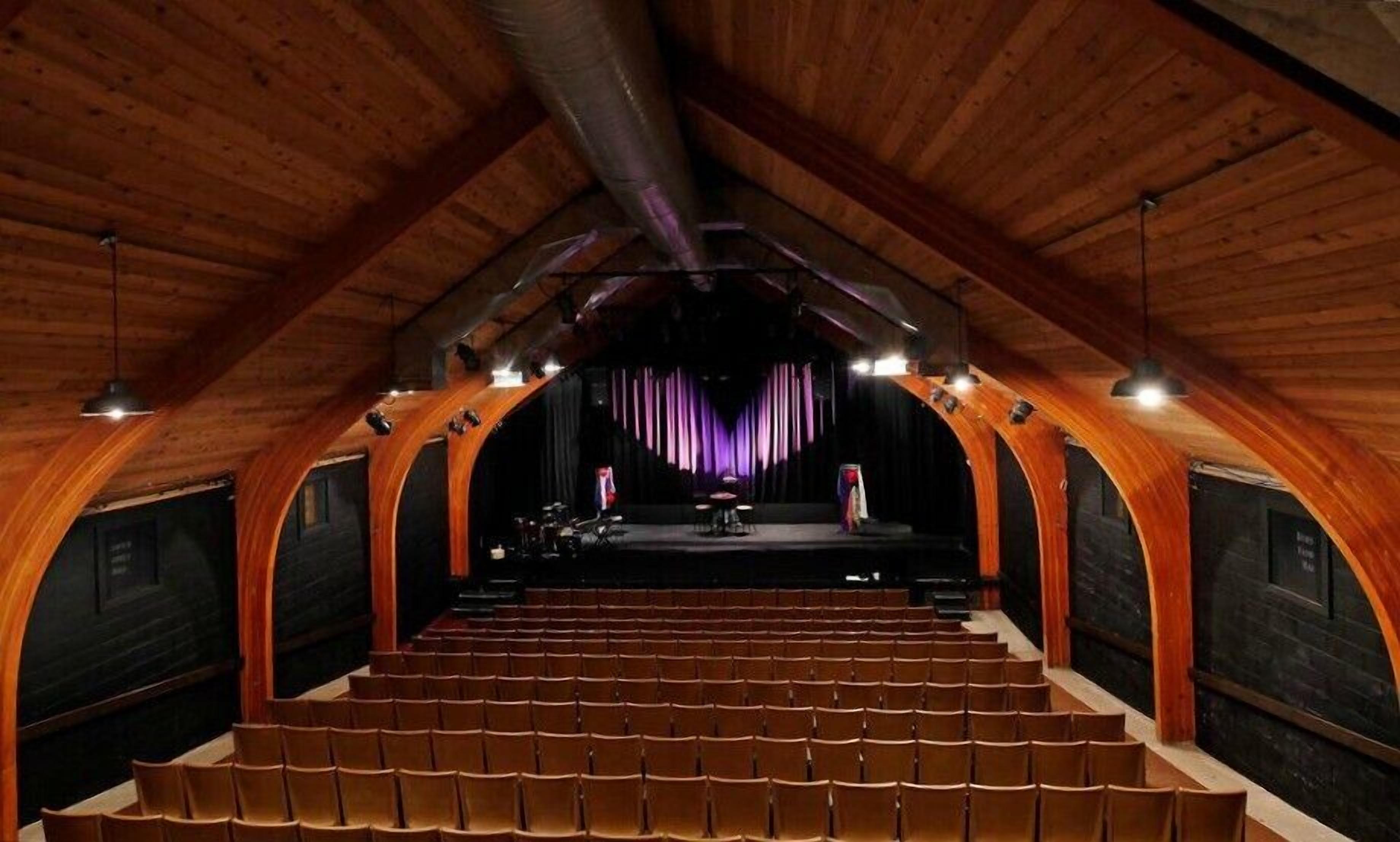 Un auditorio con asientos escalonados, un escenario con telón de fondo morado y elementos arquitectónicos de madera.