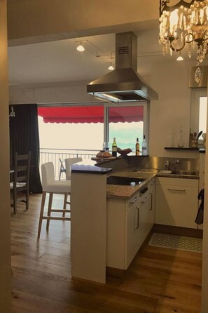Eine Küche mit einer zentralen Kochinsel, einem Essbereich mit einem Stuhl, einer Chandelüster und Blick durch ein Fenster.