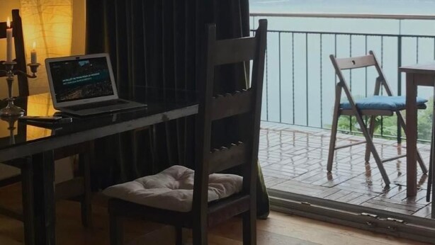 Une pièce avec un ordinateur portable posé sur une table, une chaise en bois, un lustre et une vue sur la mer depuis un balcon.