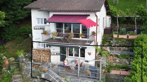 Une maison à deux étages avec un auvent rouge, des sièges à l’extérieur et des plantes en pot.