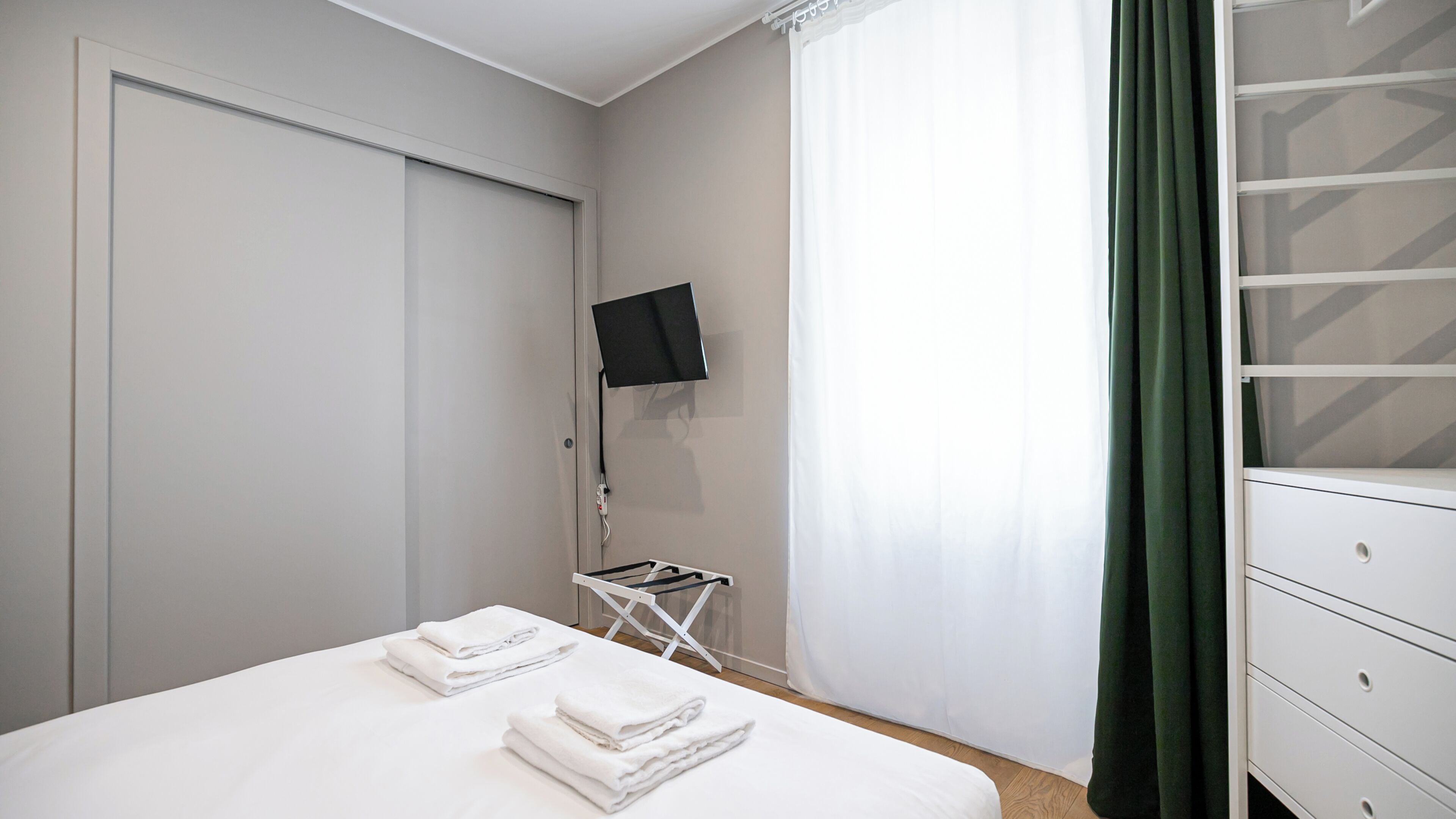 Une chambre d’hôtel avec un lit, un téléviseur fixé au mur, une petite table avec une chaise et un rideau vert.