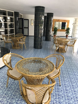 Une pièce avec une table ronde en osier et des chaises, des piliers cylindriques noirs et des carreaux de sol bleus à motifs.
