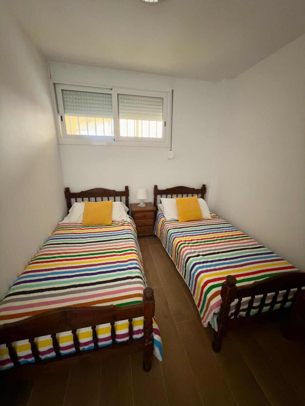 A room with two beds, each with a striped bedspread, a nightstand between them, and a window above.