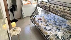 Ein Stockbett mit einem gemusterten Bettbezug, ein Standventilator, ein kleiner runder Tisch mit einer Lampe und ein Fenster mit Vorhängen.