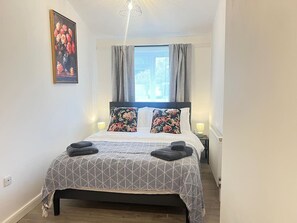 Ein Schlafzimmer mit Bett, Kissen, einem Bild, einem Fenster mit Vorhängen und einer Nachttischlampe.