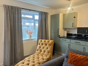 Ein Zimmer mit Fenster, einem gepolsterten Sessel, einer Couch mit orangefarbenem Kissen, einer Küchenzeile mit Schränken und einer Pendelleuchte.