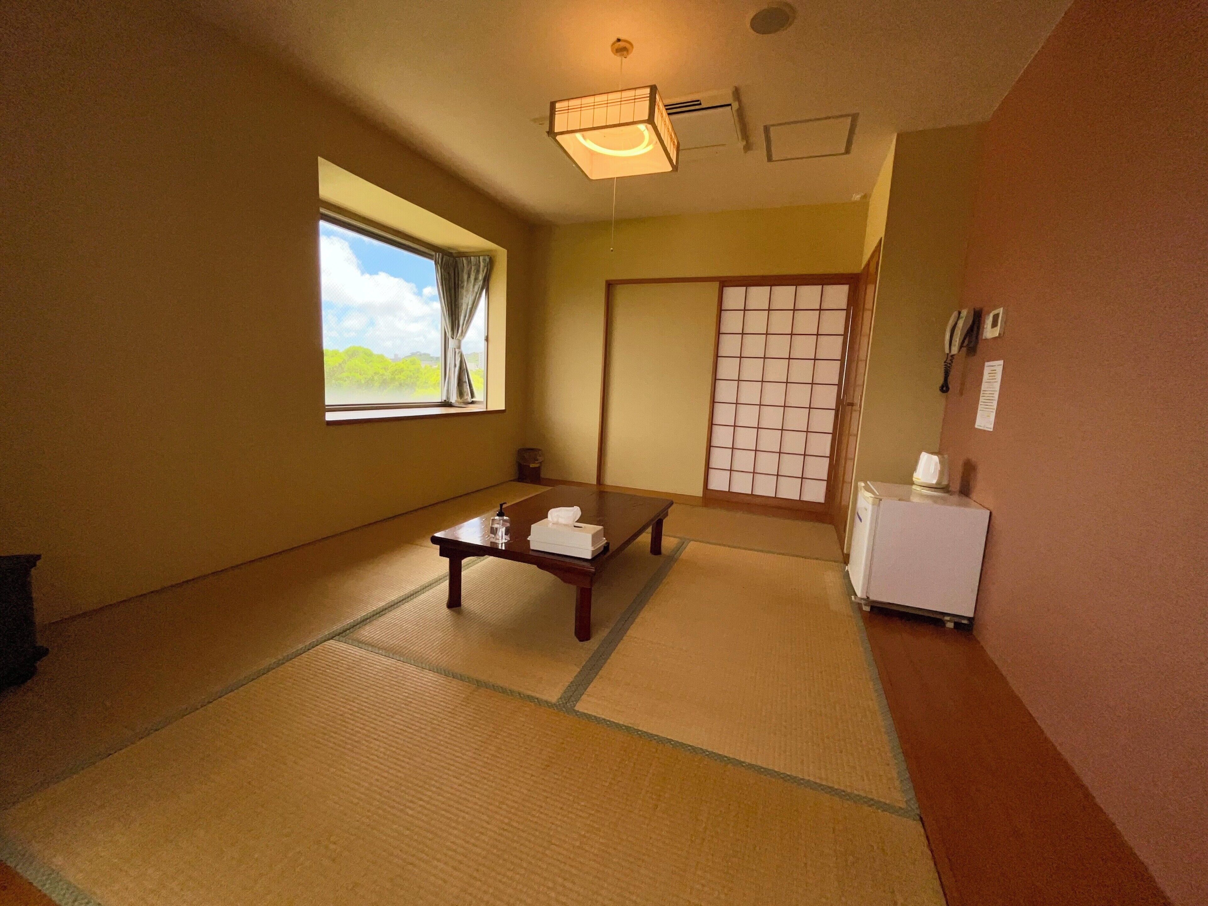 Una habitación con esteras de tatami, una mesita baja con una toalla, un refrigerador pequeño y un divisor de puertas correderas shoji.