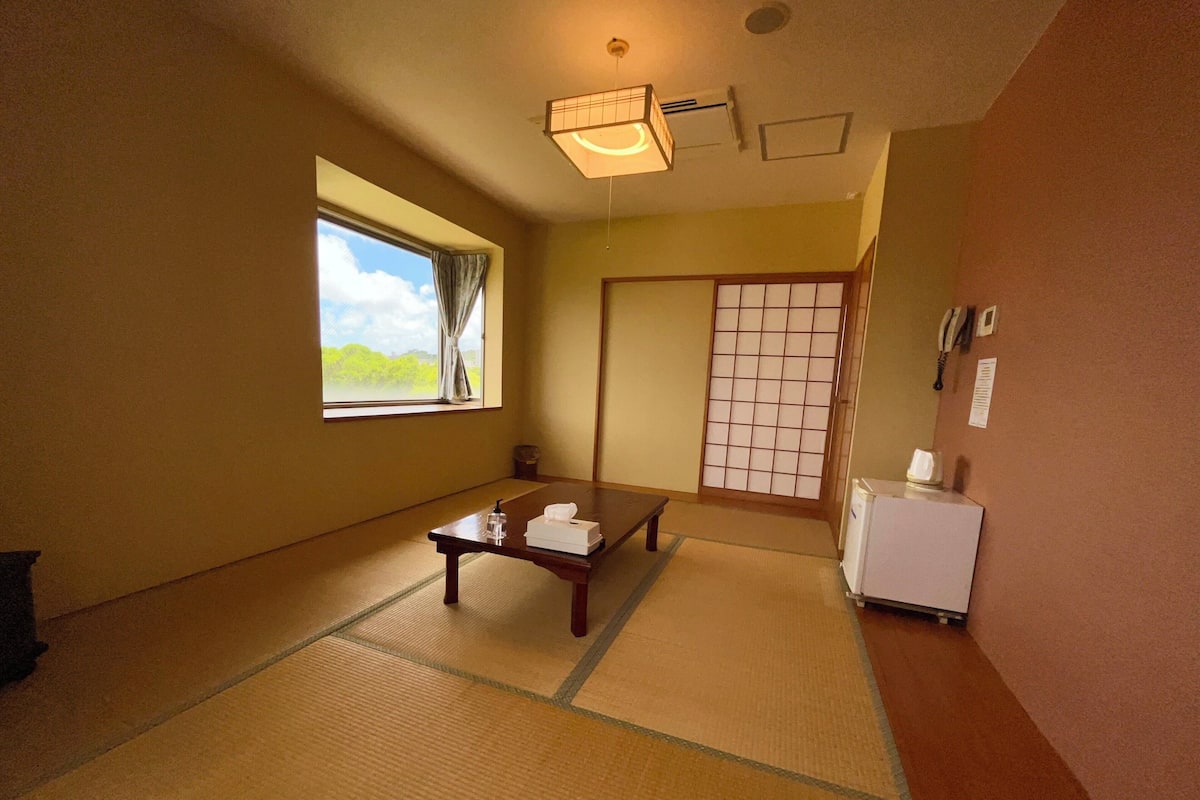 A room with tatami mats, a low table with a towel, a small refrigerator, and a shoji screen.