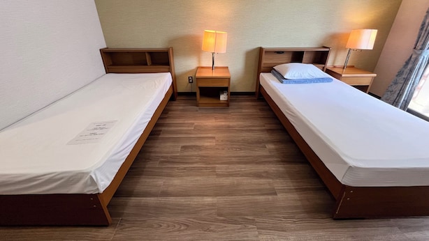 A room with two single beds, wooden headboards, bedside tables with lamps, a small shelf, and a window with curtains. - Enagic OKinawaInternational Youth Hostel (Naha)