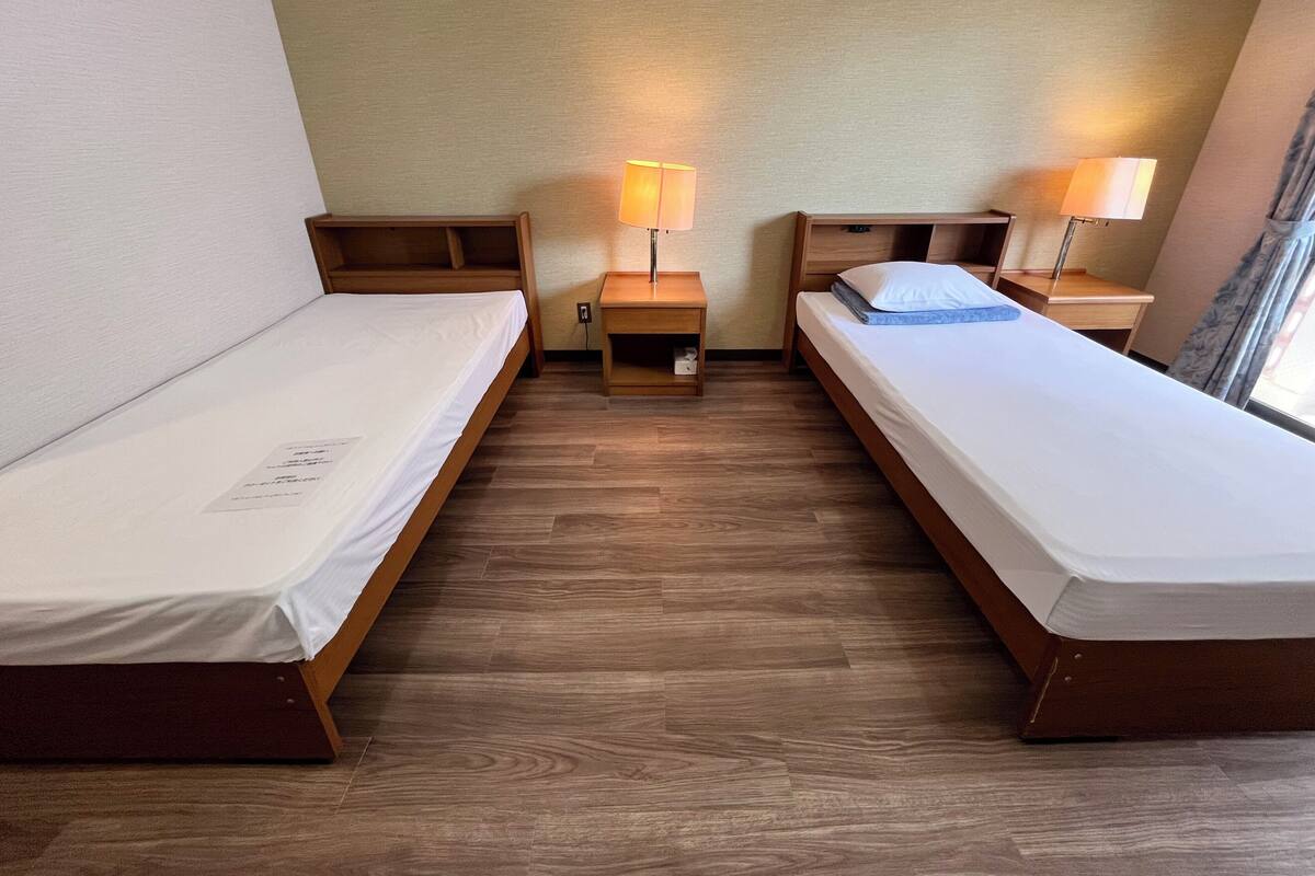 A room with two single beds, wooden headboards, bedside tables with lamps, a small shelf, and a window with curtains.
