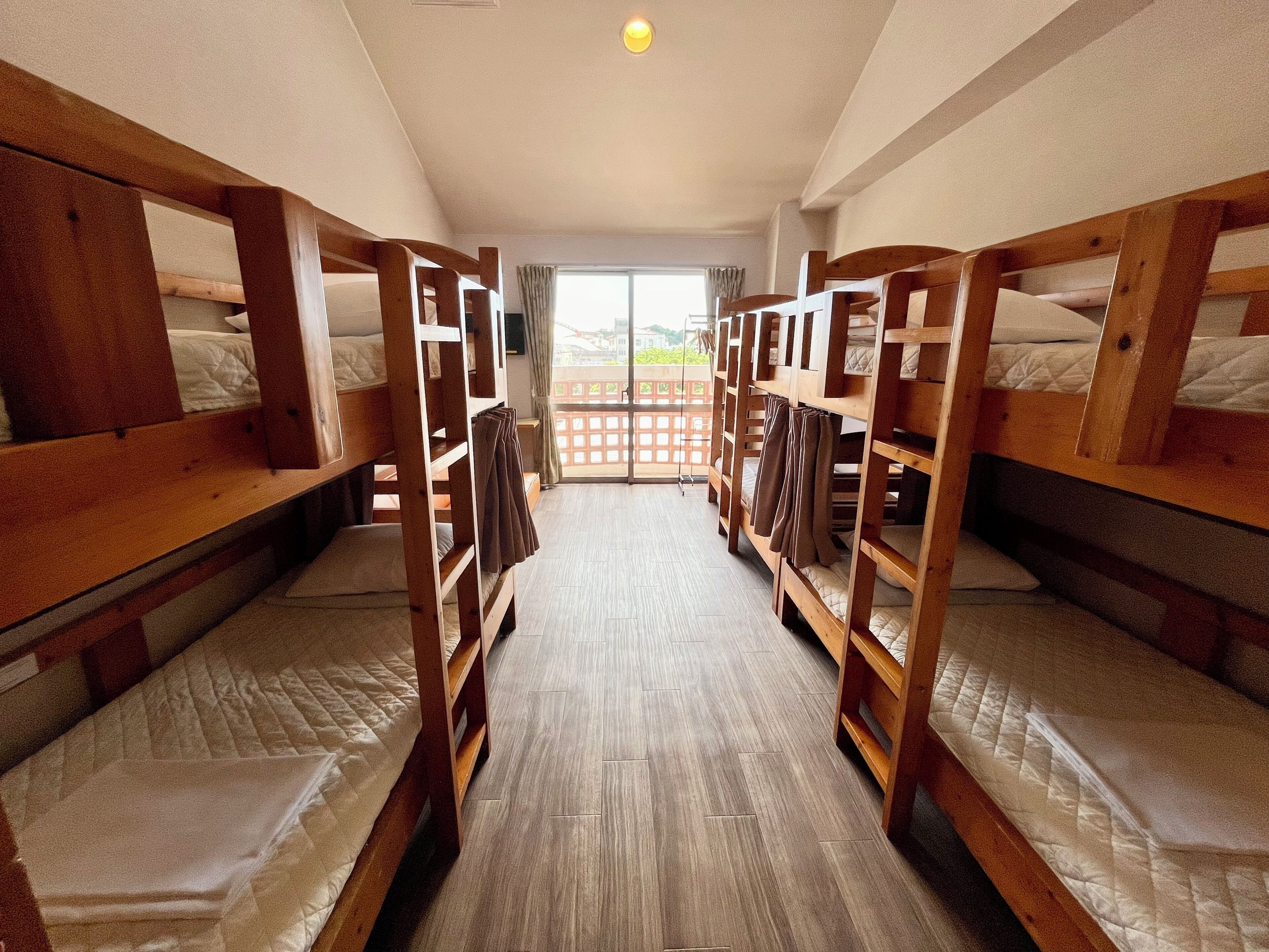 Una habitación con literas alineadas, suelo de madera y vista por una ventana.