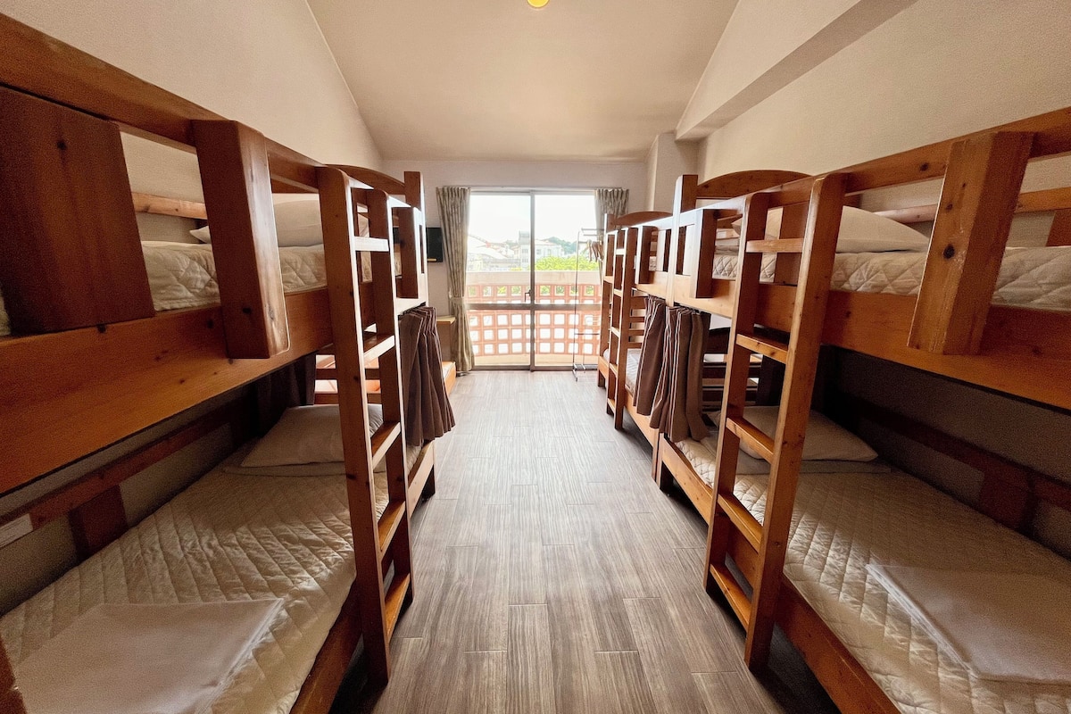 A room with bunk beds arranged in a row, a wooden floor, and a view through a window.