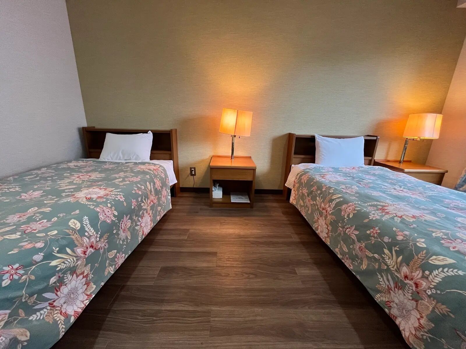 Una habitación de hotel con dos camas individuales, cabeceros de madera, mesitas de noche con lámparas y suelo de madera.