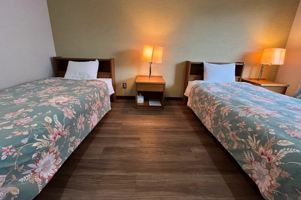 A hotel room with two single beds, wooden headboards, nightstands with lamps, and a wooden floor.