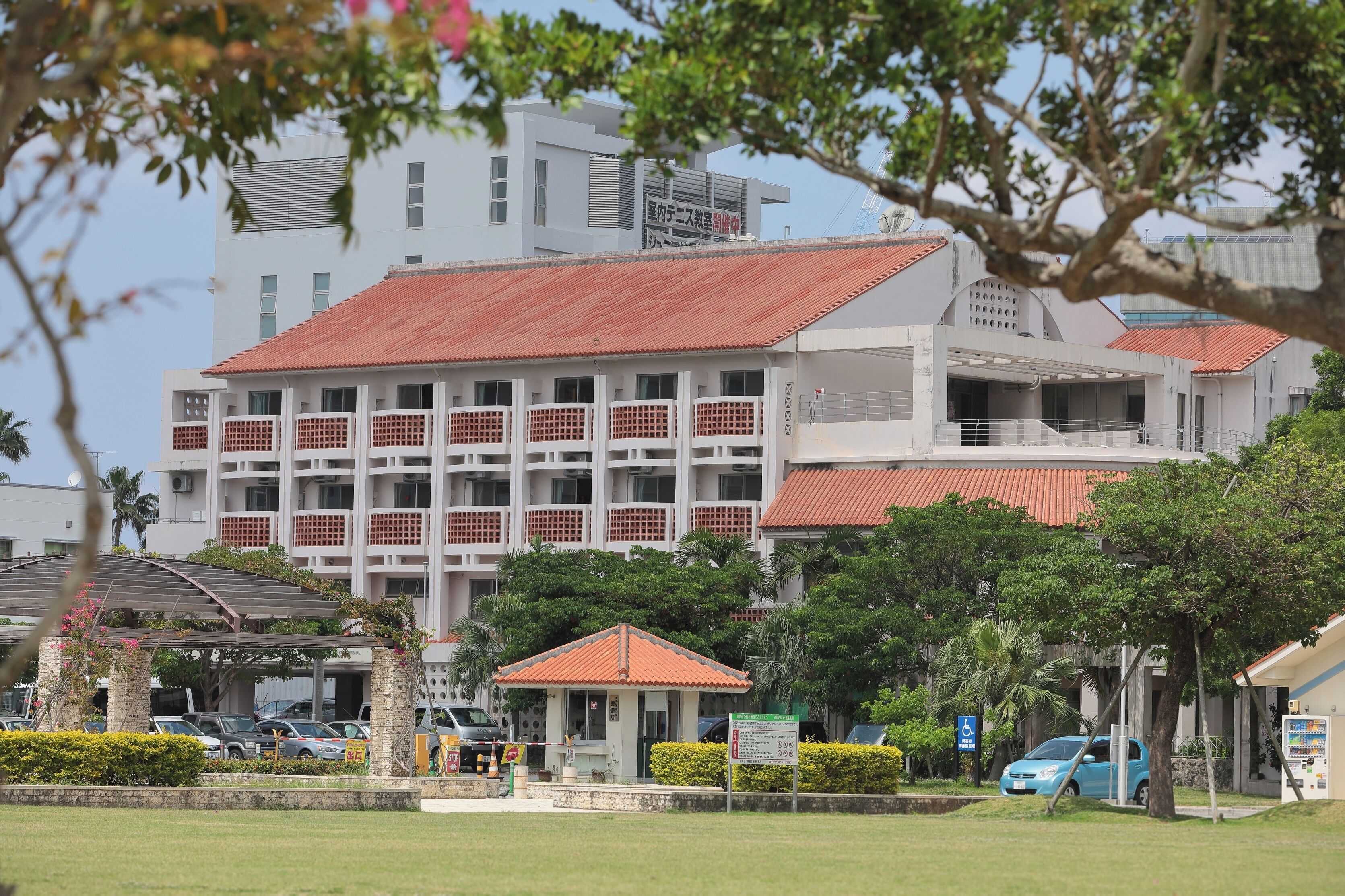 A multi-story building with a red-tiled roof, a covered pavilion, and a parking area with cars.