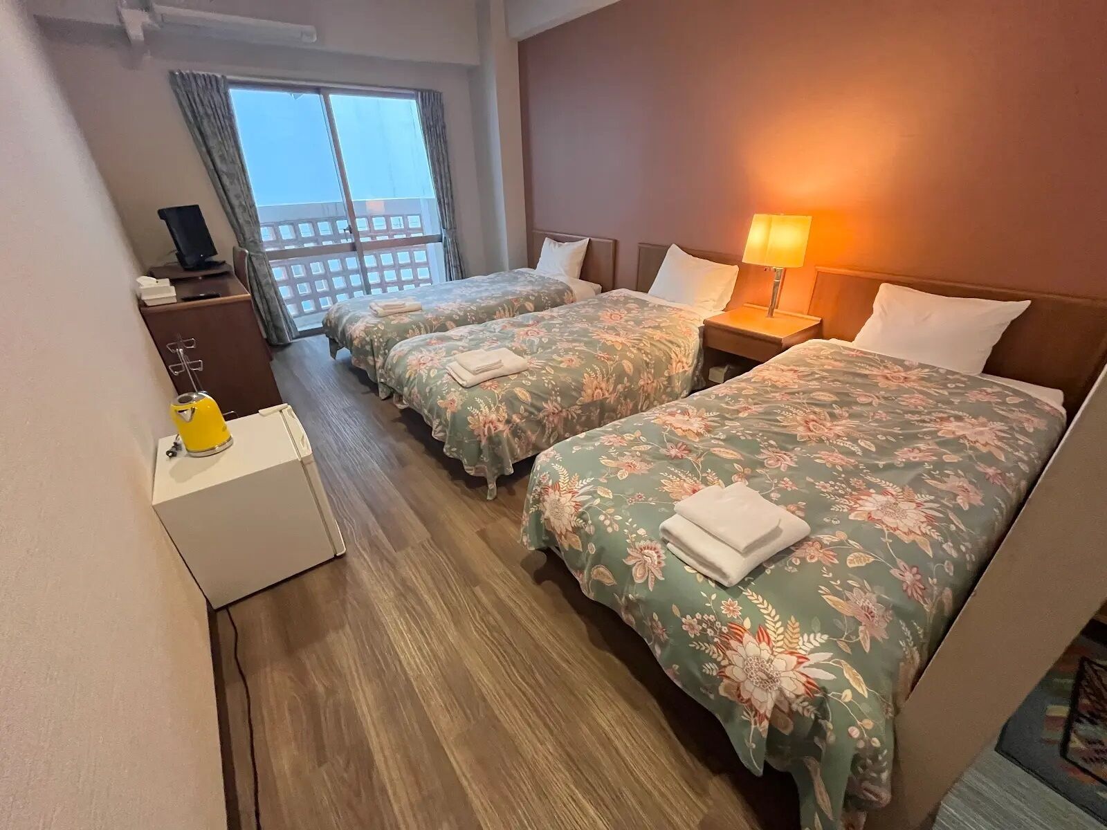 Habitación de hotel con dos camas, un escritorio con lámpara, una mesita con hervidor y vistas al exterior.