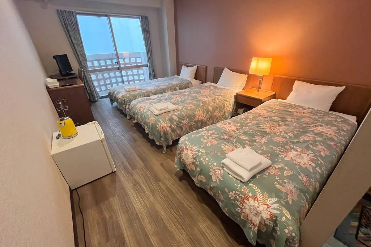 A hotel room with two beds, a desk with a lamp, a small table with a kettle, and a view of the outside.