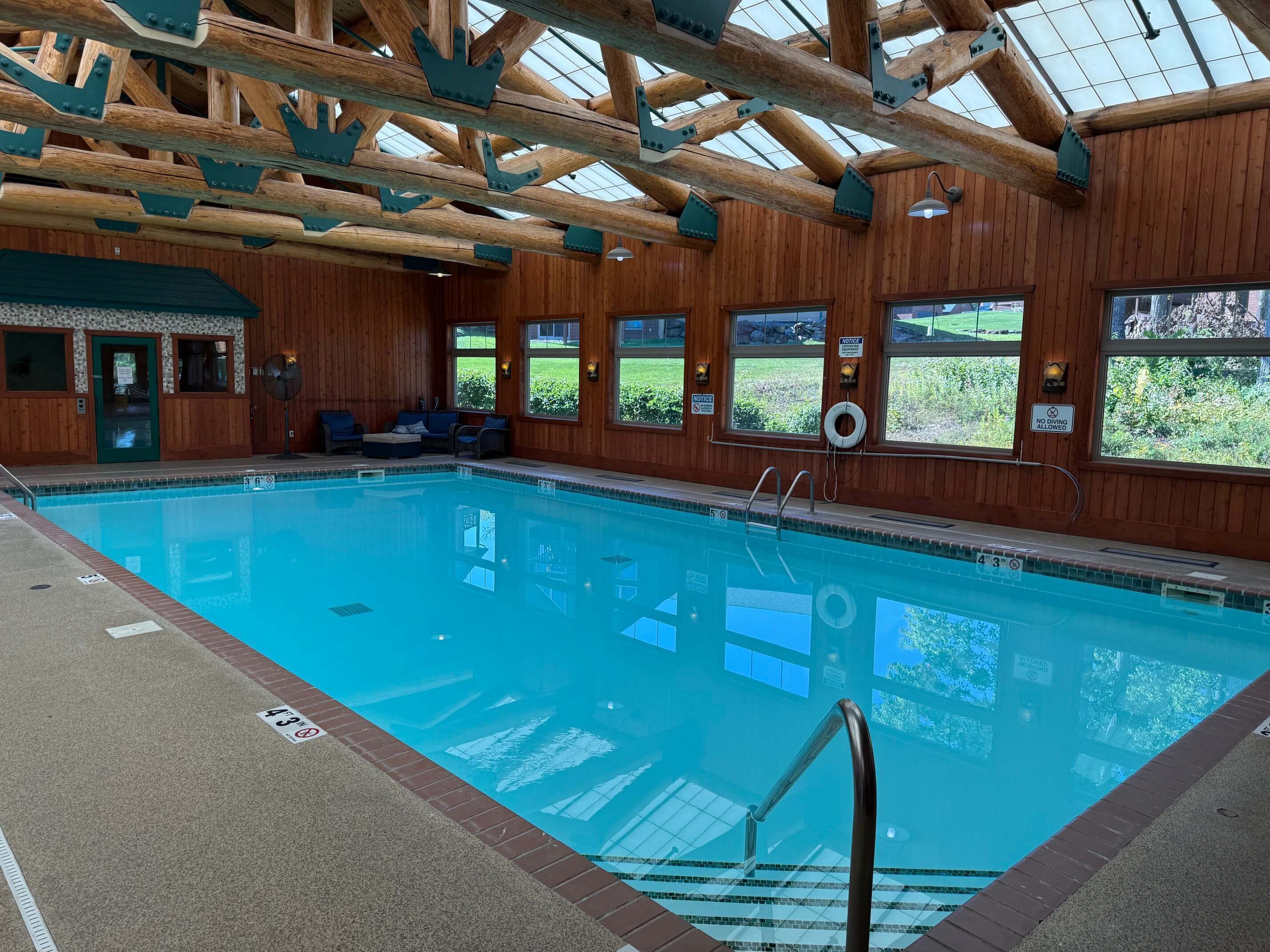 Uma piscina interior com vigas de madeira, uma cadeira de salva-vidas e vista para o exterior através de grandes janelas.