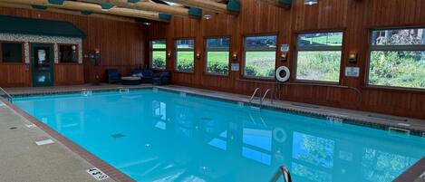 Uma piscina interior com vigas de madeira, uma cadeira de salva-vidas e vista para o exterior através de grandes janelas.