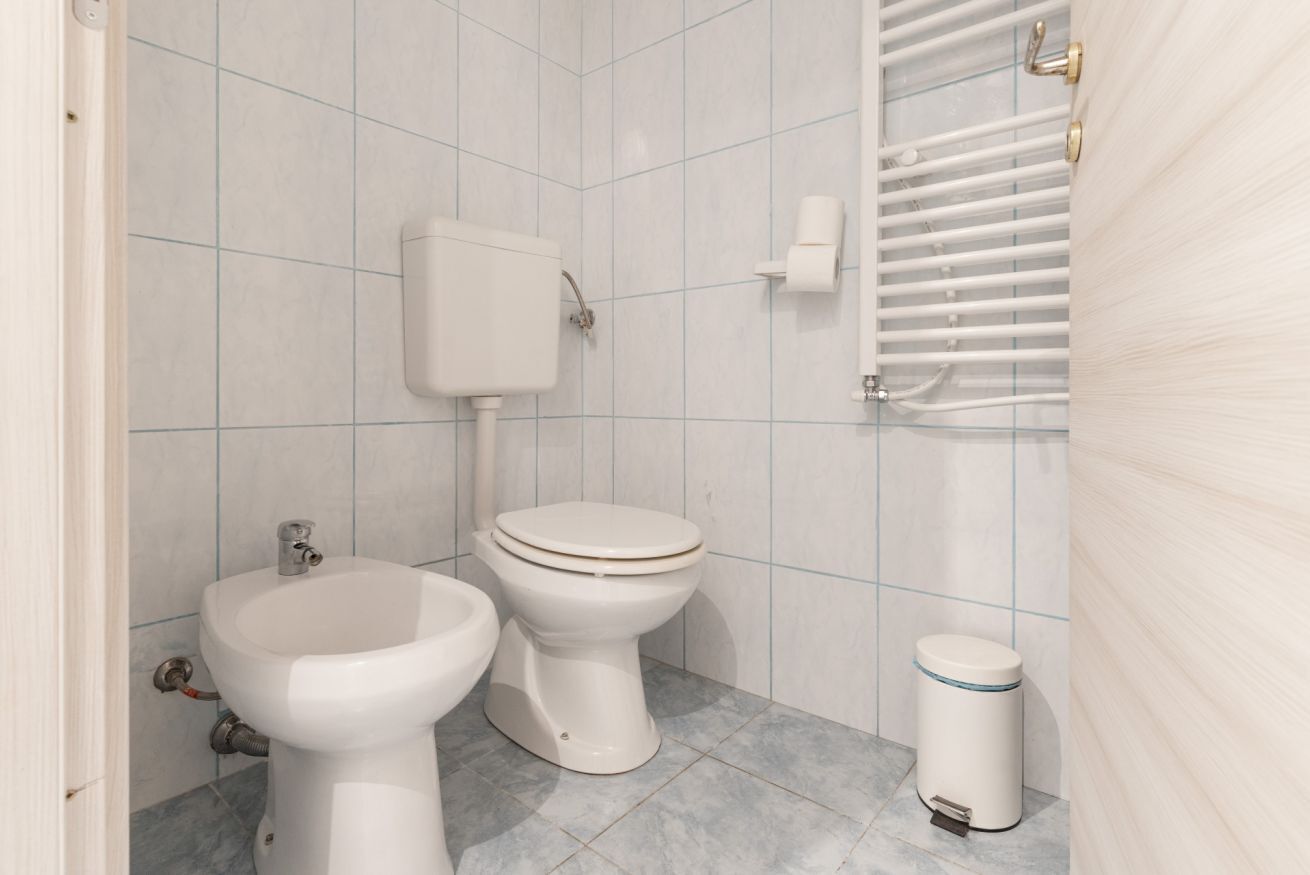 A bathroom with two toilets, a bidet, a toilet paper holder, a trash bin, and a towel rack.